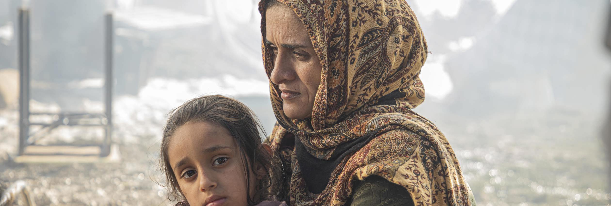 A refugee mother and daughter sit against a backdrop of rubble.