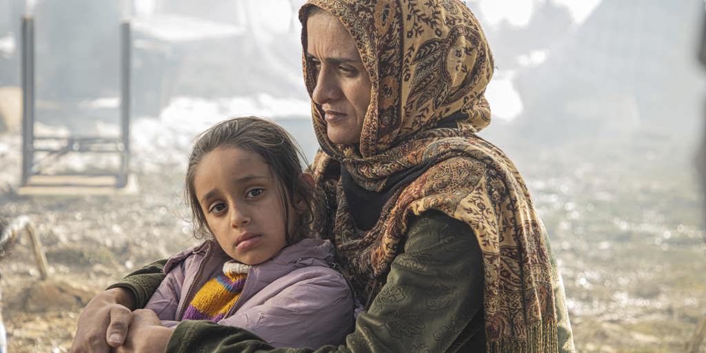A refugee mother and daughter sit against a backdrop of rubble.