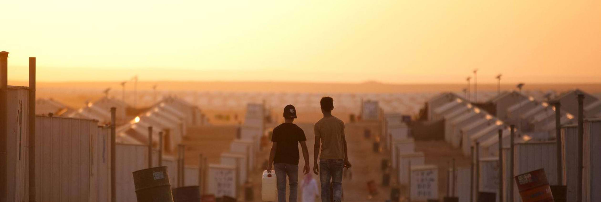 Two young refugee boys walking through a village of temporary tent accommodation