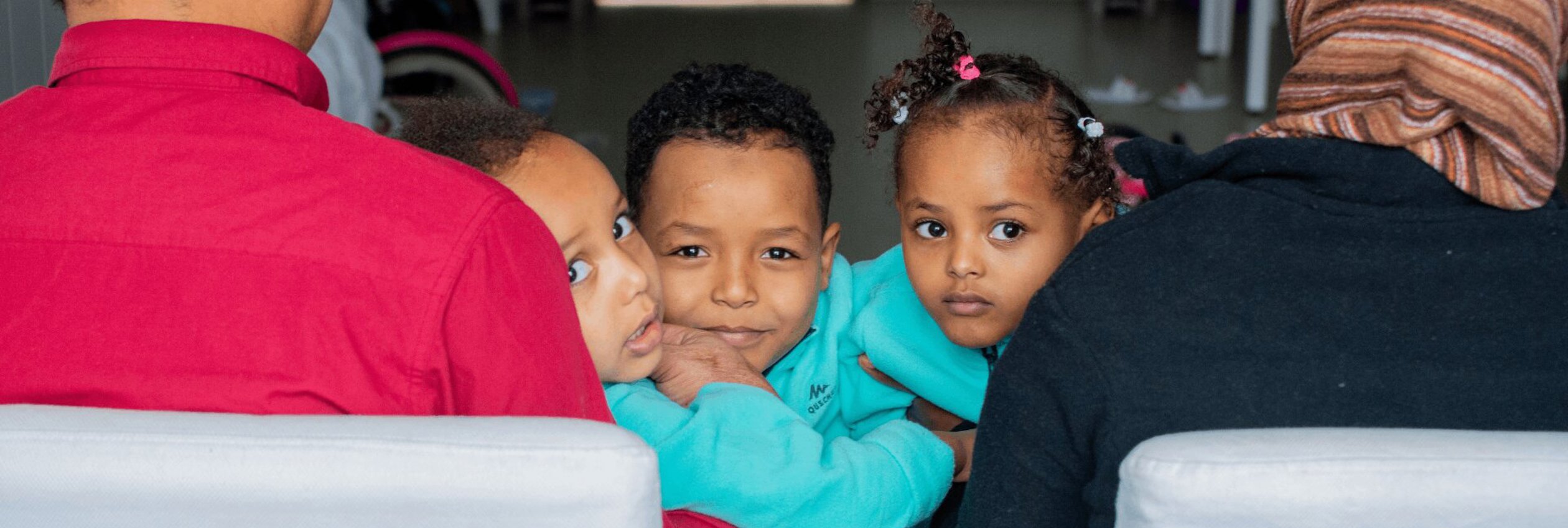 Three Eritrean children peek at the camera from their parents' laps