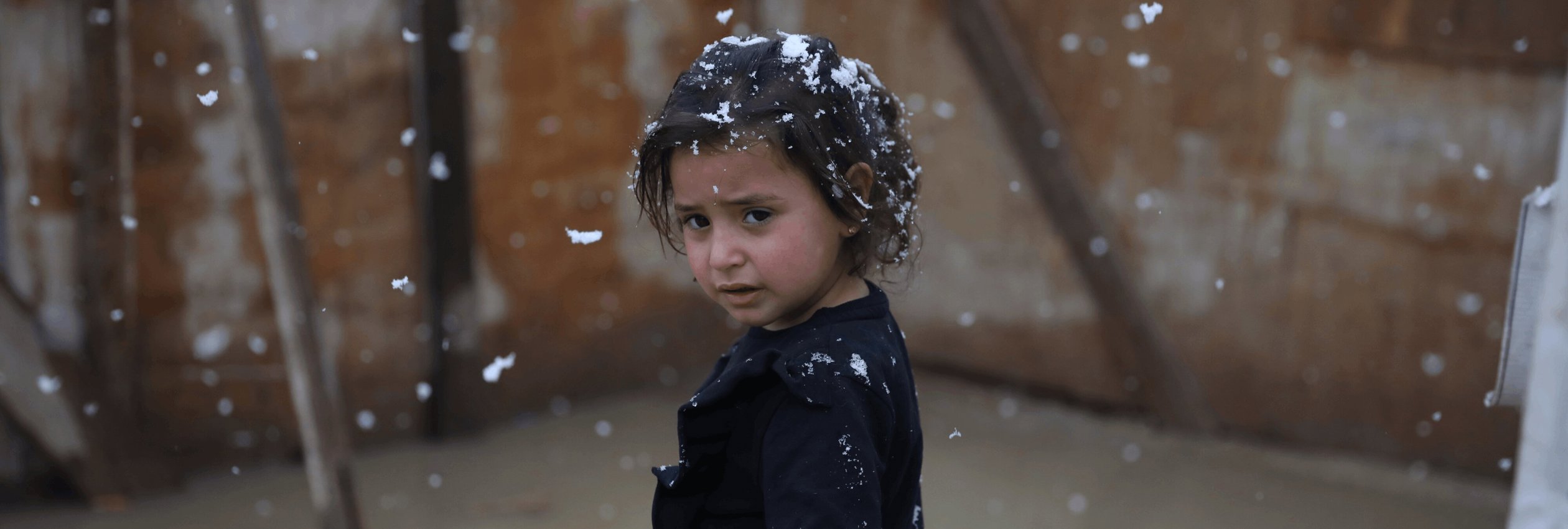 Aisha, a young Syrian girl, stands outside her family's tent in Lebanon while snow falls around her.